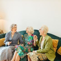 Three people chatting on a teal sofa in a bright living room with a table lamp nearby