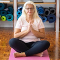 Woman sitting cross-legged on a pink yoga mat in a studio, hands together in prayer pose.
