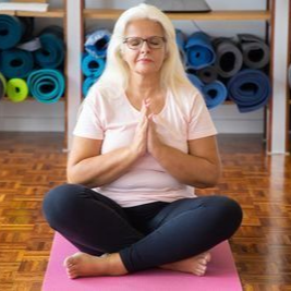 Woman sitting cross-legged on a pink yoga mat in a studio, hands together in prayer pose.