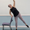 Woman stretching sideways beside a chair in a studio, wearing black top and gray leggings.