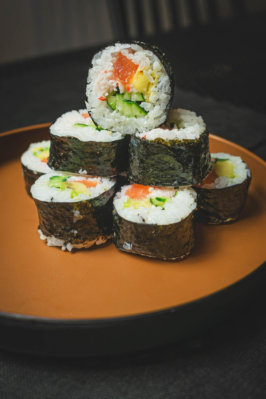Involtini di sushi impilati su un piatto marrone, con salmone, avocado e cetriolo.