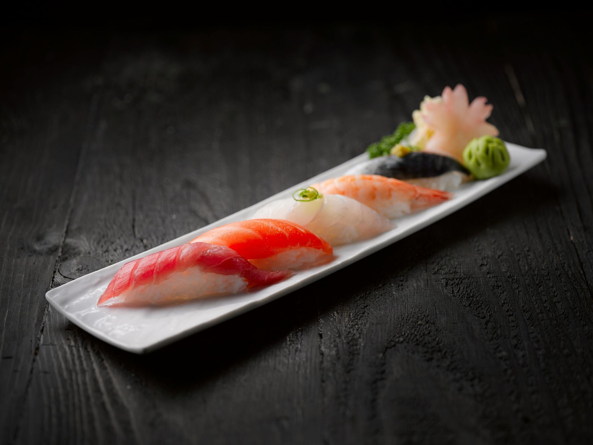 Sushi nigiri disposto su un piatto rettangolare bianco sopra una superficie di legno scuro.