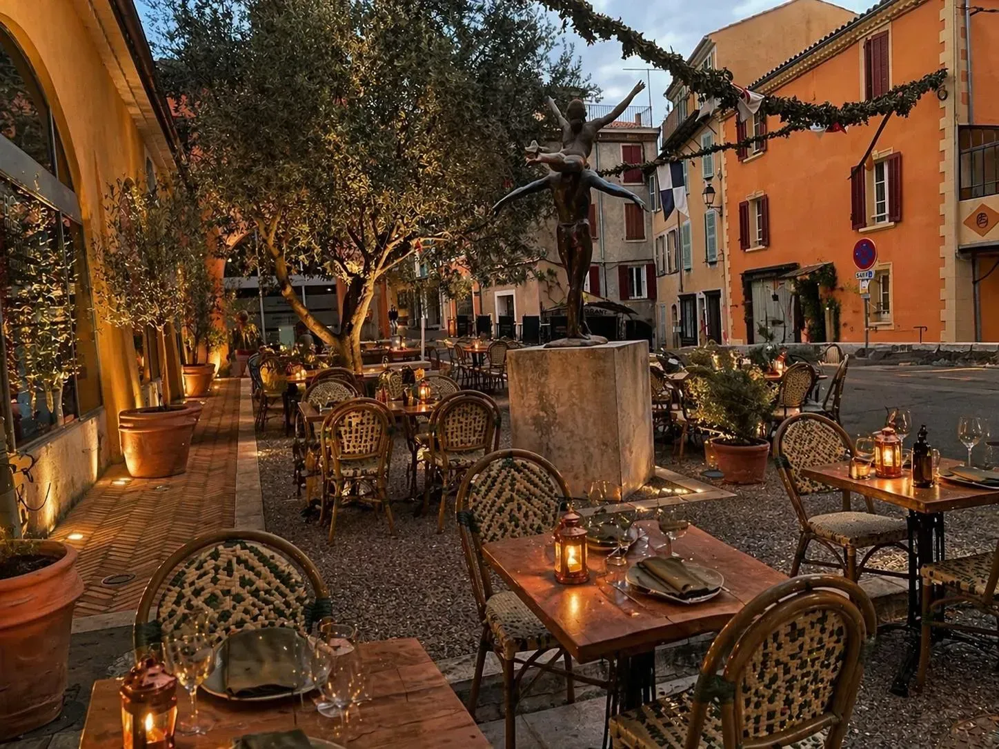 Des tables de café en terrasse accueillantes, éclairées par une lumière chaleureuse, dans une rue étroite bordée de bâtiments colorés.
