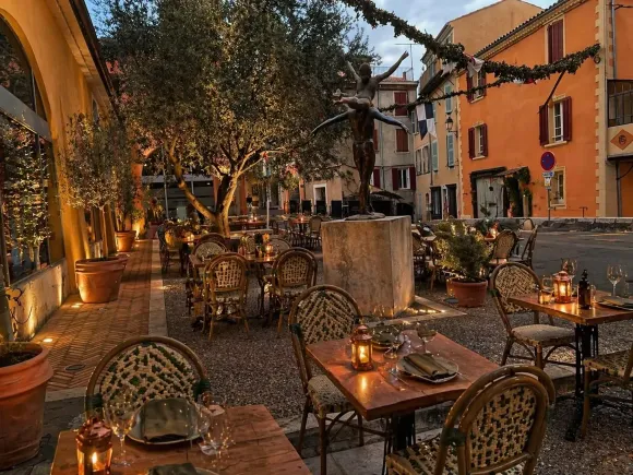 Terrasse de café extérieur accueillante au crépuscule, avec chaises en osier, tables et lumières chaudes sous les arbres.