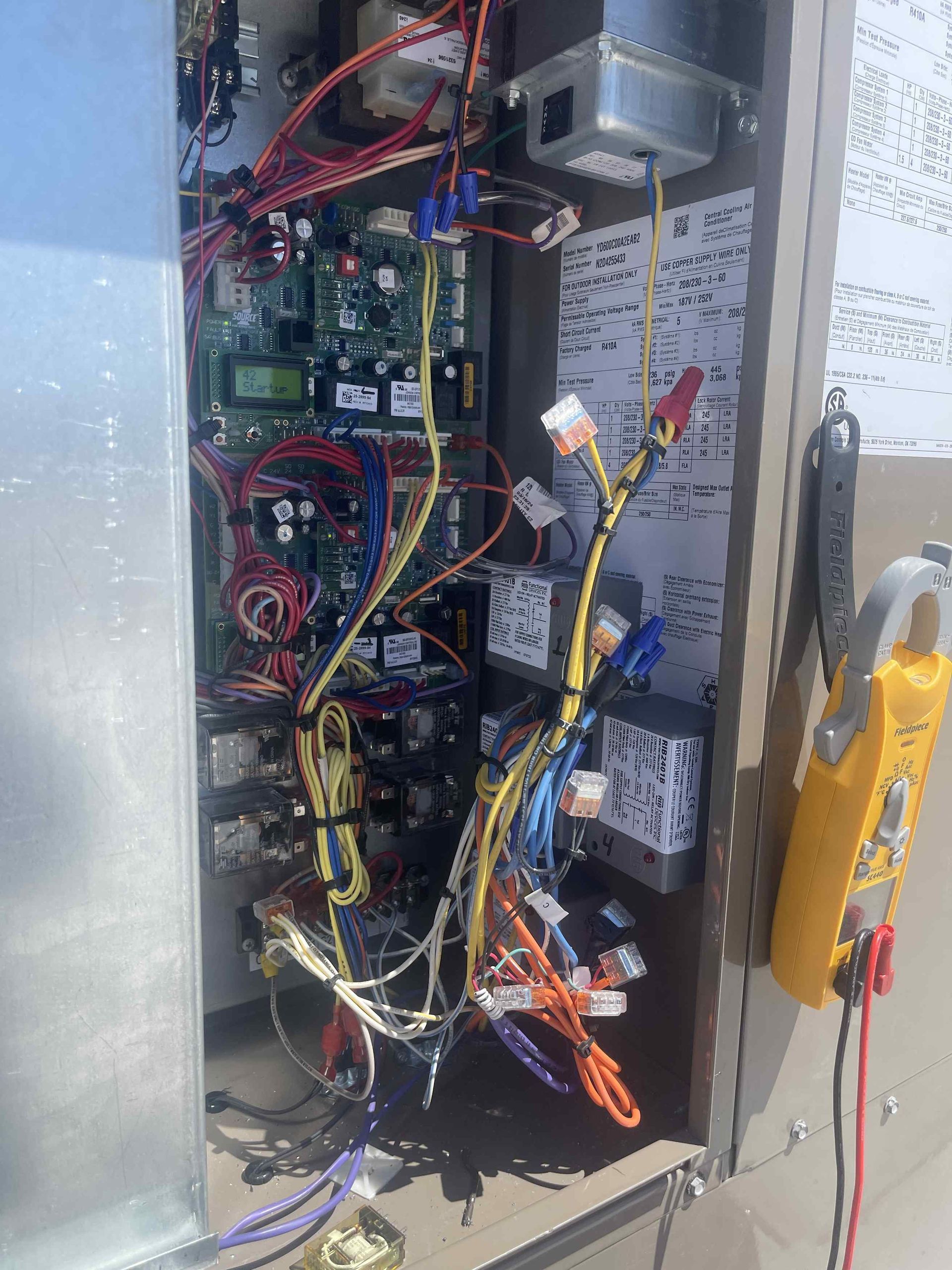 Open electrical panel on a HVAC system with wires and circuit boards, plus a clamp meter.