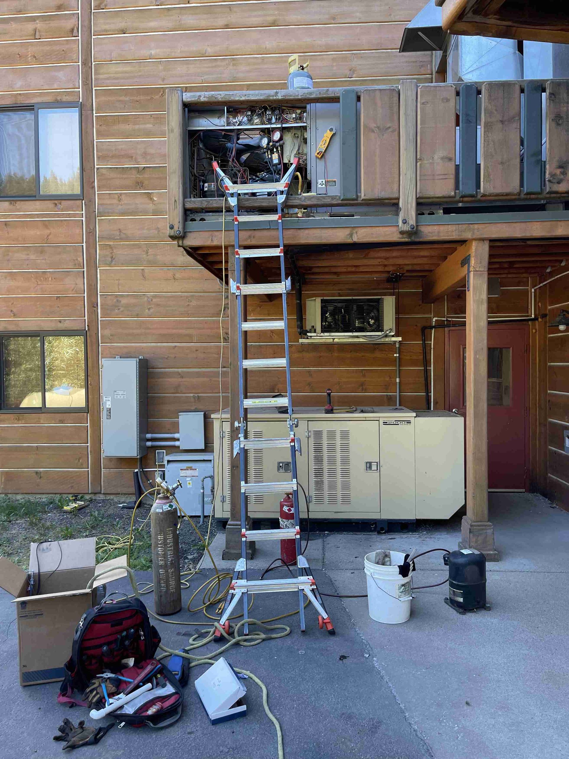 HVAC unit repair on wooden building. Ladder, tools, and equipment on the ground.