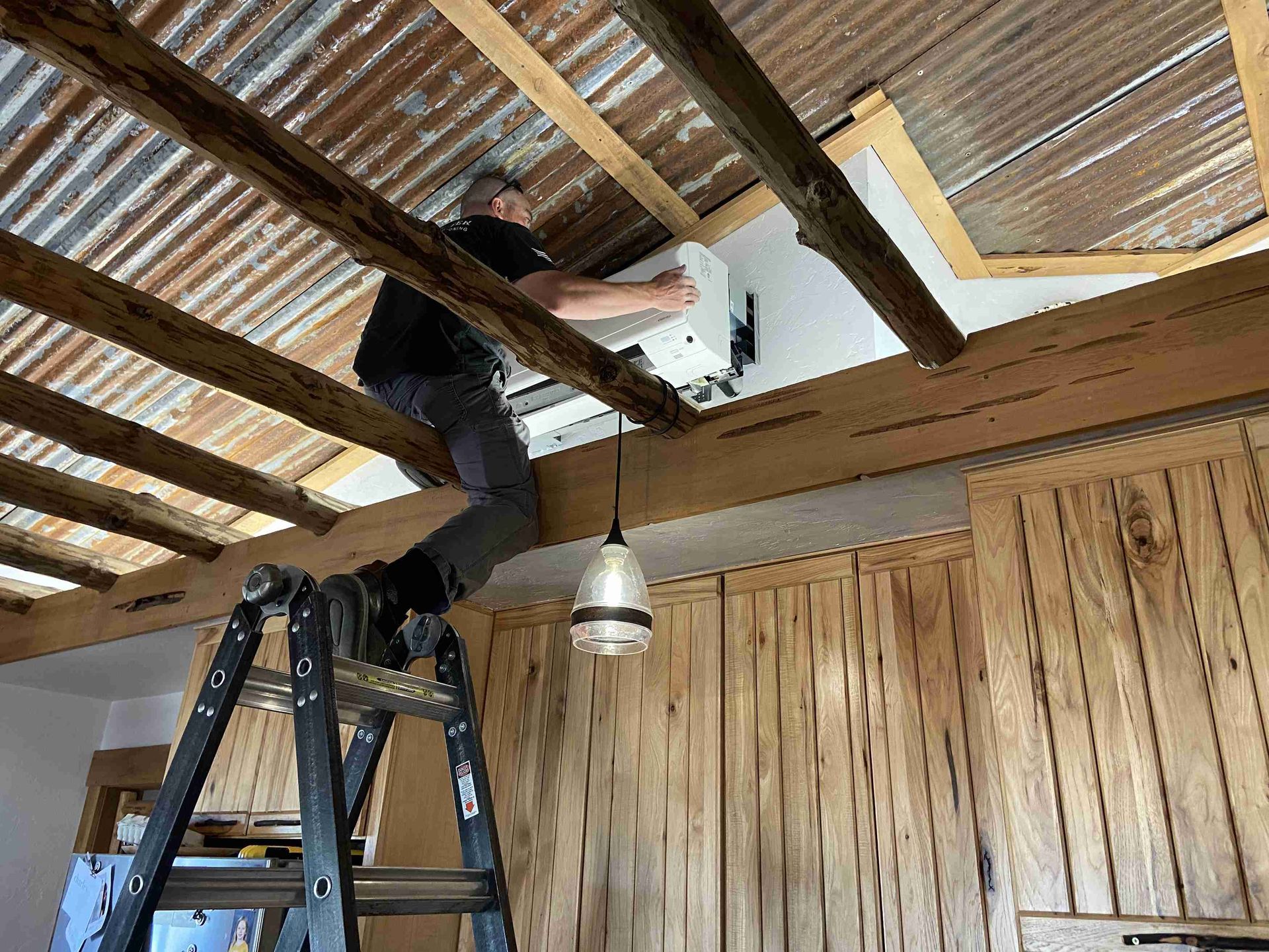 Man on ladder installs object on white wall, wooden beams and corrugated metal ceiling.