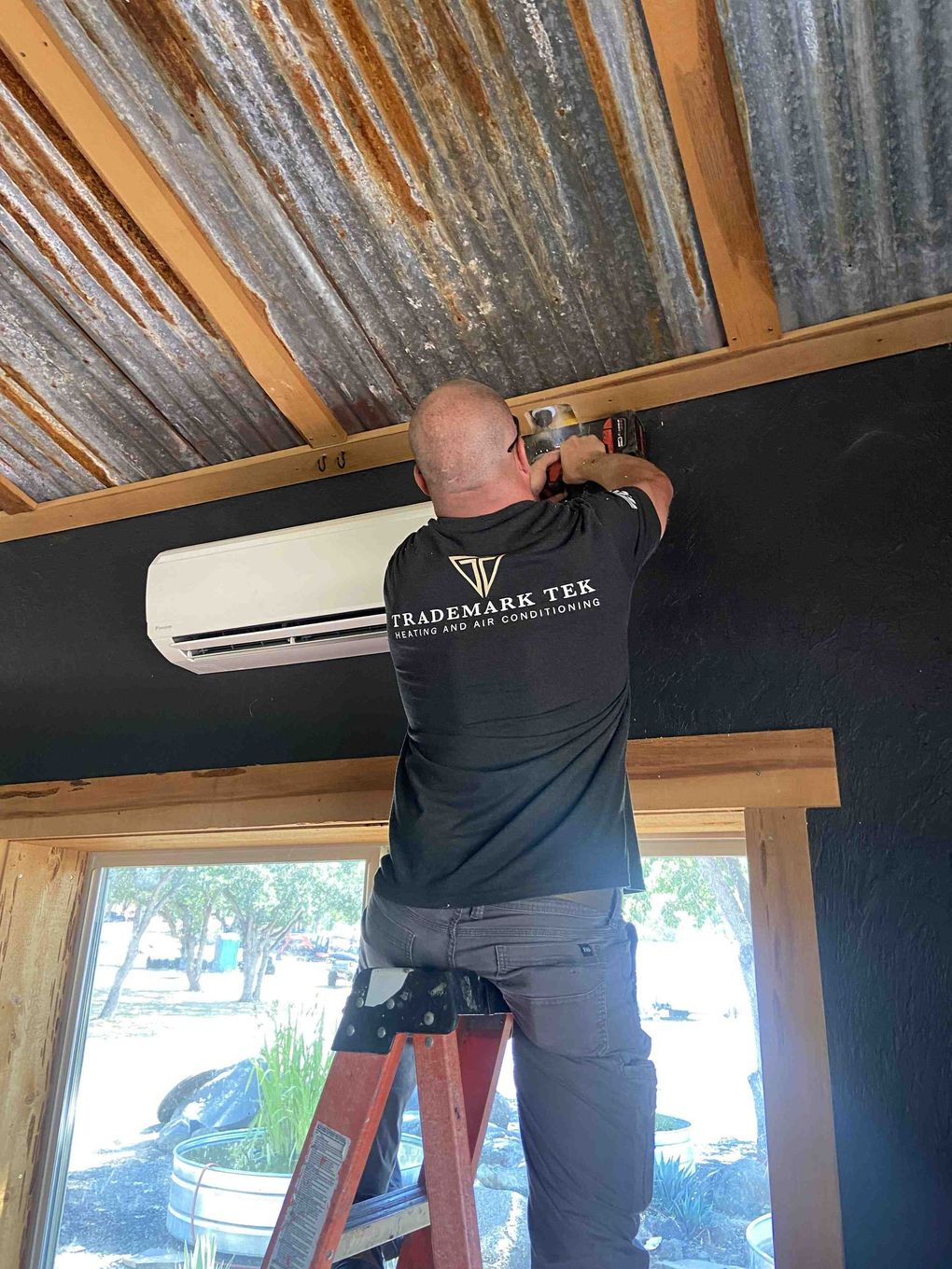 Man on a ladder installing an AC unit on a black wall, under a corrugated metal ceiling in a Medford, Oregon home.