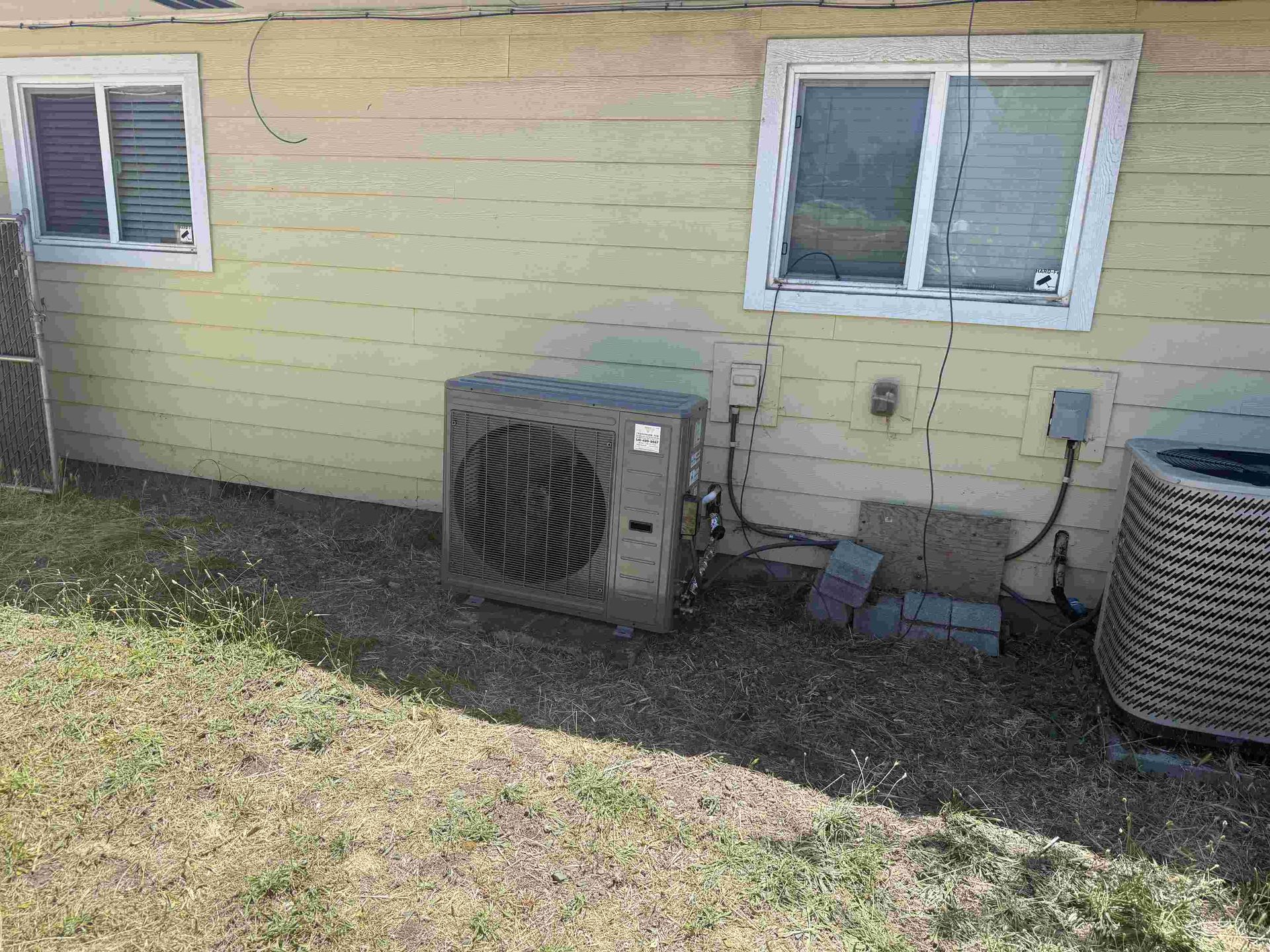 An air conditioning unit next to a house with windows and yellow siding on dry grass.