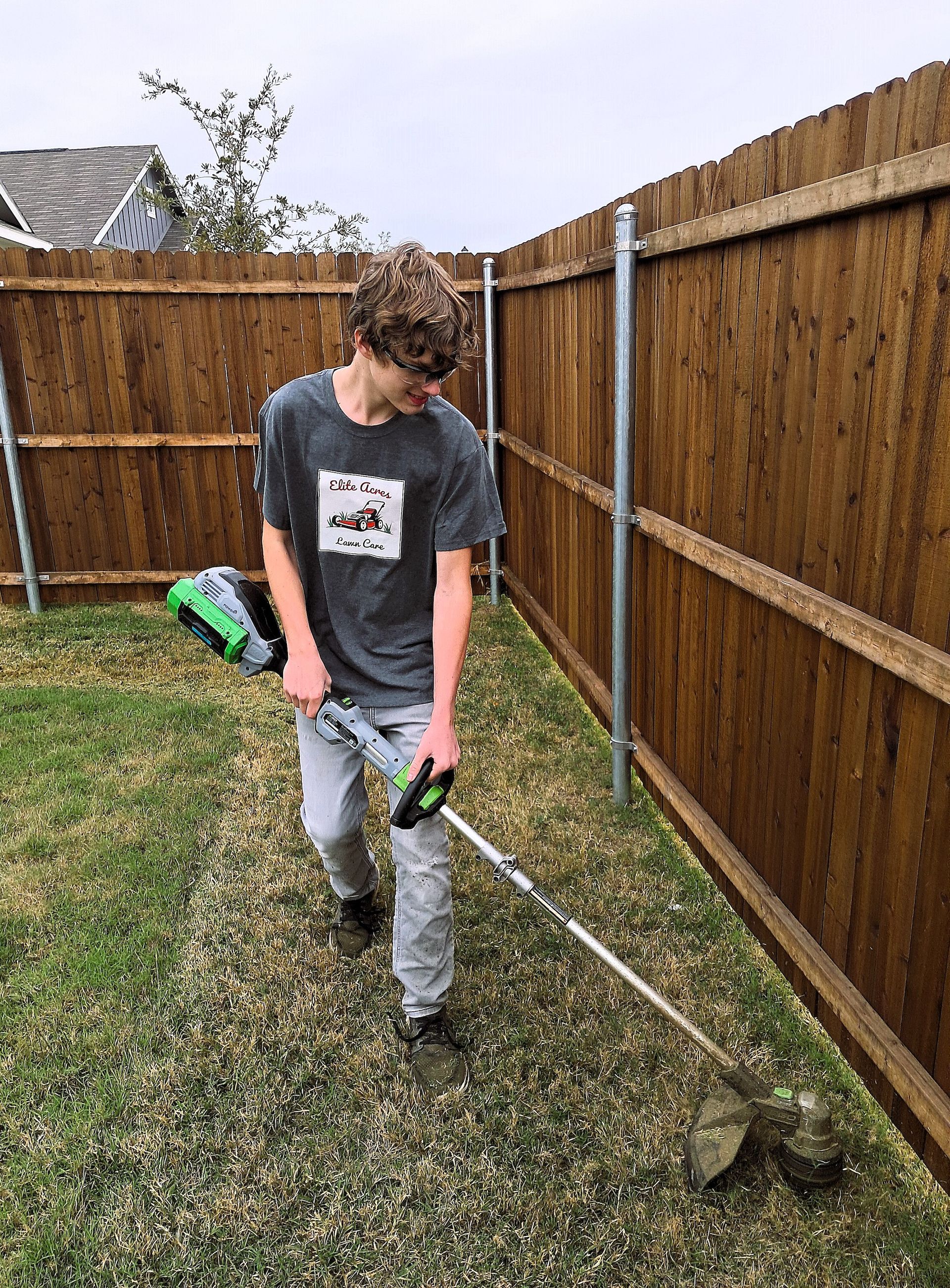 A young man is using a weed eater in a backyard next to a wooden fence.