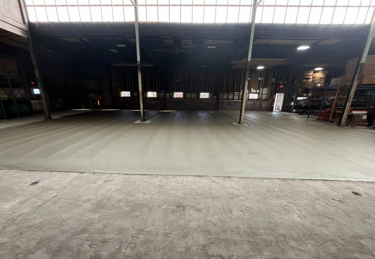 Empty warehouse interior with a large concrete floor and open bay doors under skylights