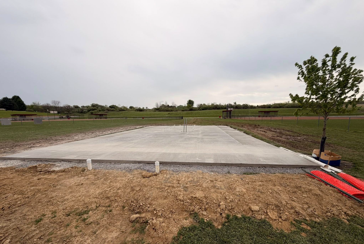 Fresh concrete slab under construction in a grassy field with a small tree and tools nearby