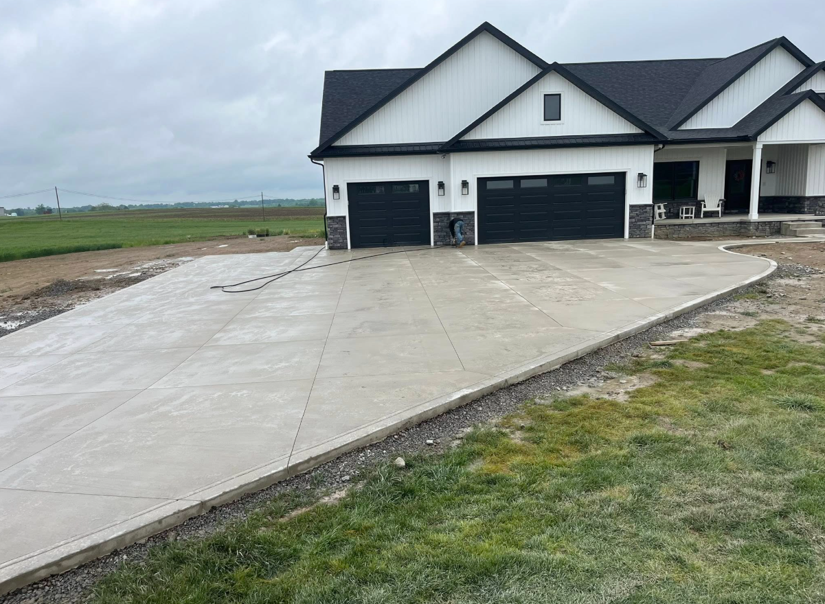 New concrete driveway leading to a white house with black garage doors on a cloudy day
