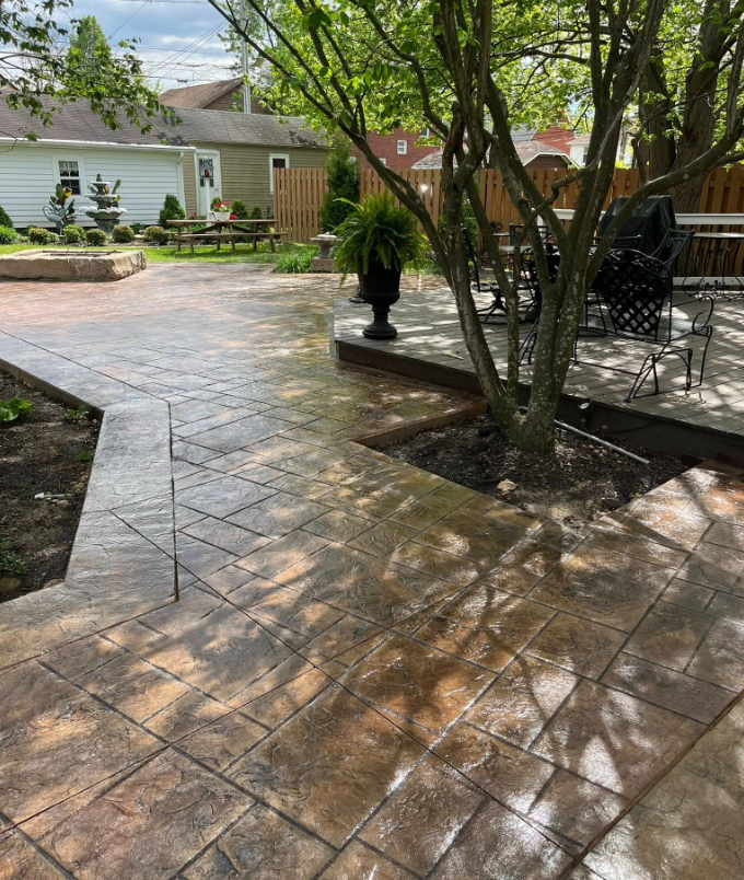 Backyard patio with stamped concrete, trees, and outdoor seating beside a house.