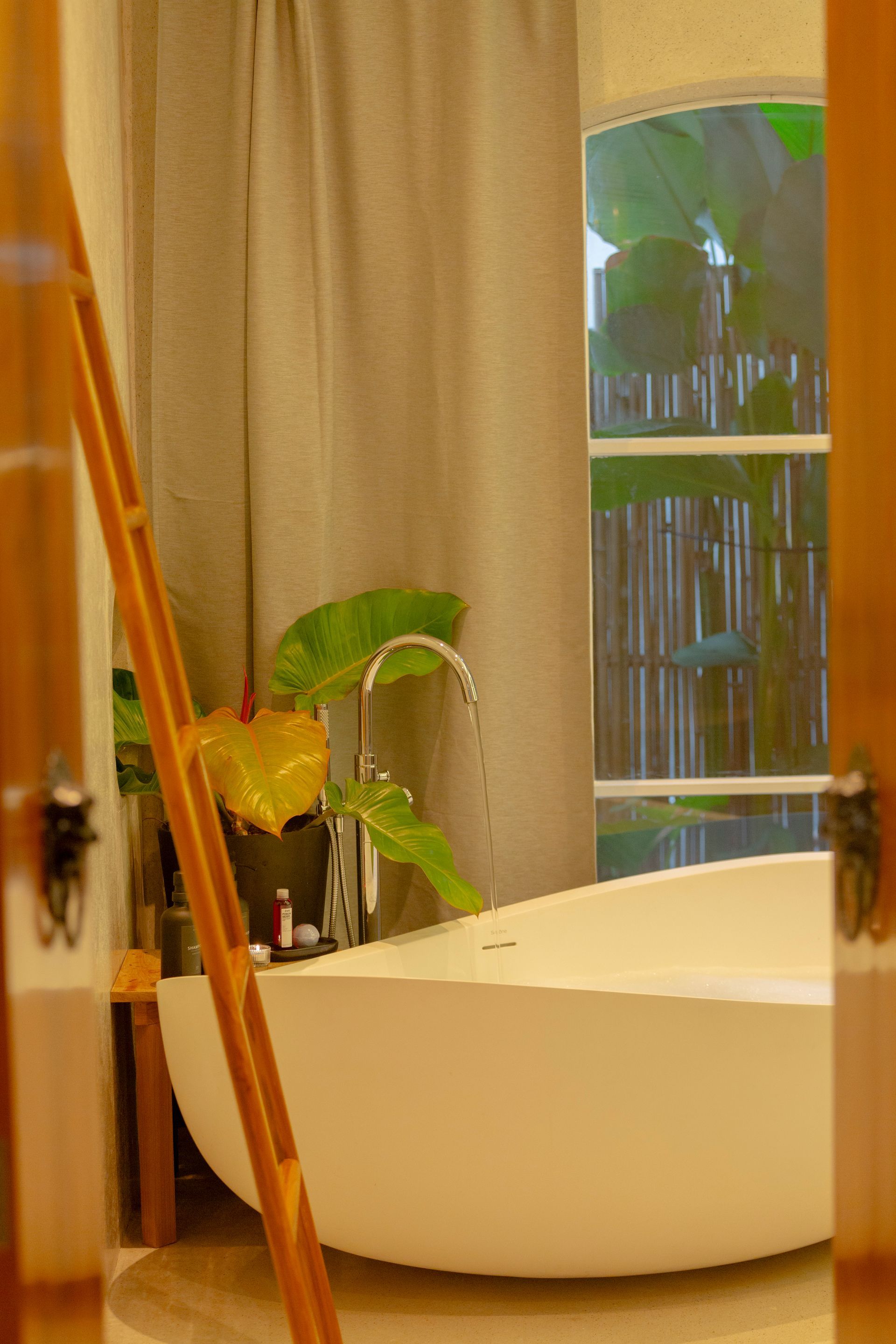 Bathroom with white tub, faucet, plant, window with bamboo screen, tan curtain, and light.