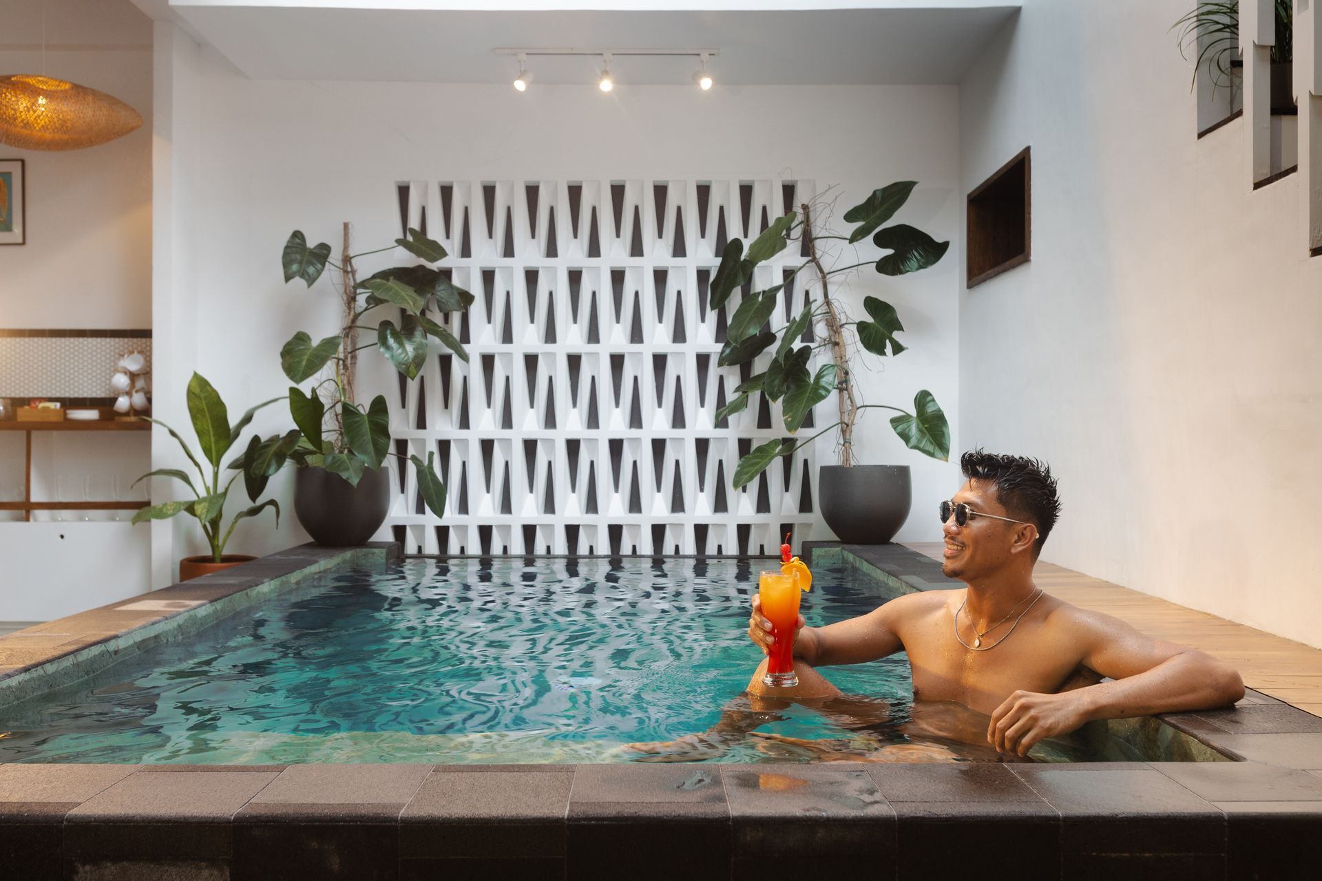 Man in sunglasses in a pool, holding a cocktail. Plants and patterned wall in a white-walled room.