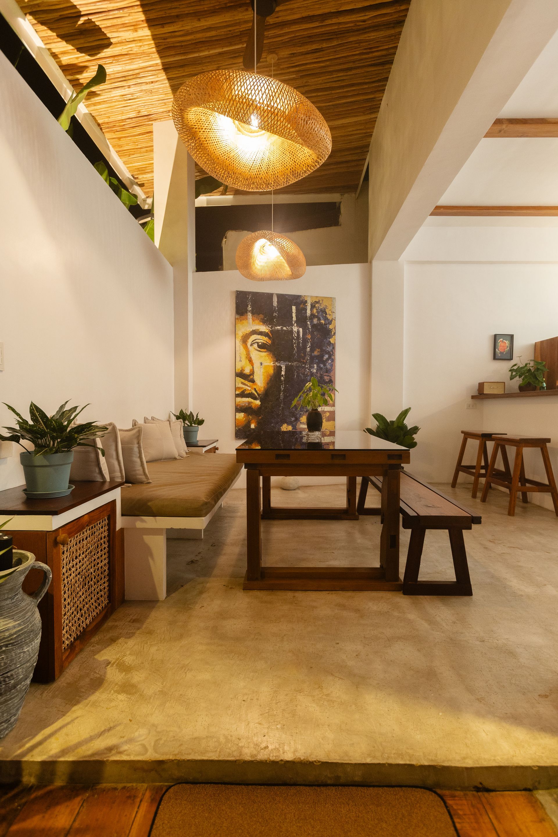 Dining area with wooden table, bench, and hanging light fixtures. Buddha art on the wall.