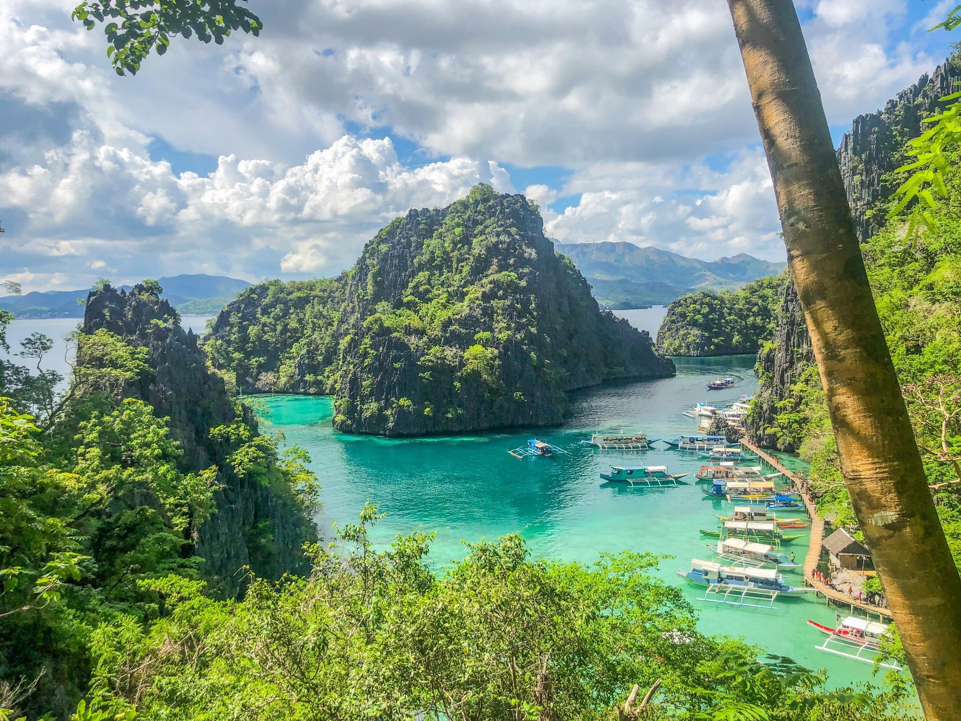 Turquoise water surrounded by lush, green cliffs and boats, under a cloudy sky.
