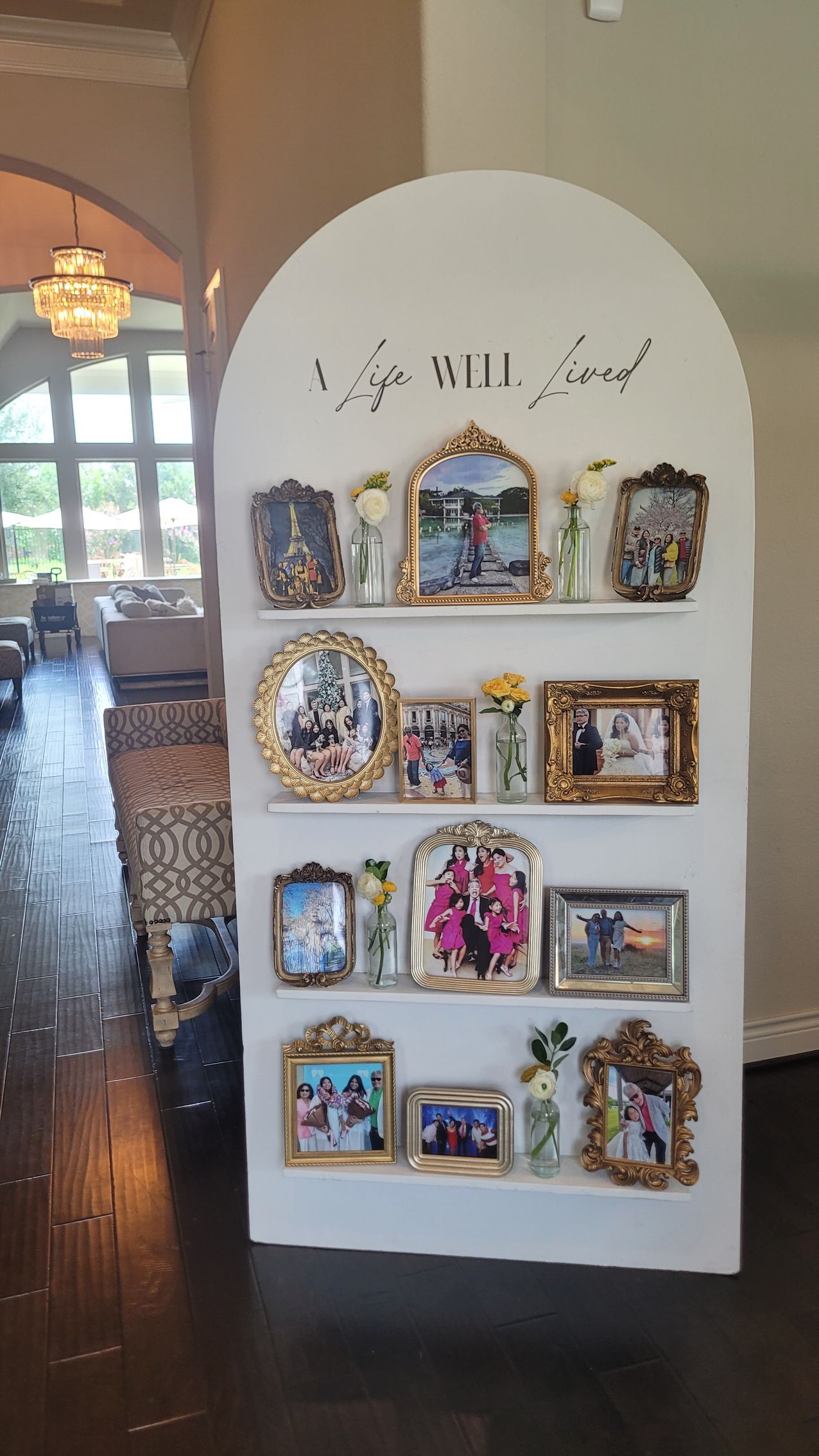 White arch display shelf with A Life Well Lived memorial photo display in vintage gold frames and yellow floral bud vases for a celebration of life decor setup