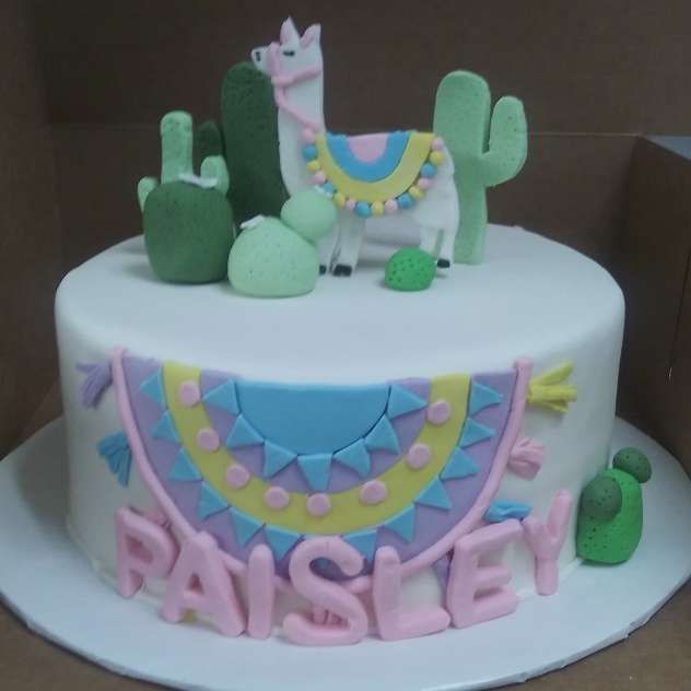 White cake with pastel cactus decorations and a llama topper, with “RAISHEY” in pink letters on the front.