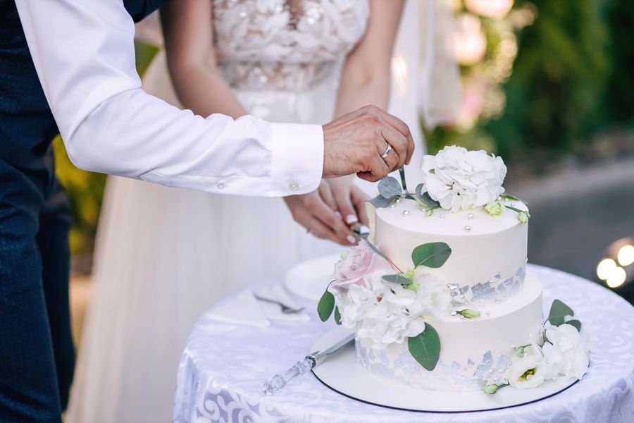 Hands cutting a white wedding cake decorated with flowers on a table outdoors