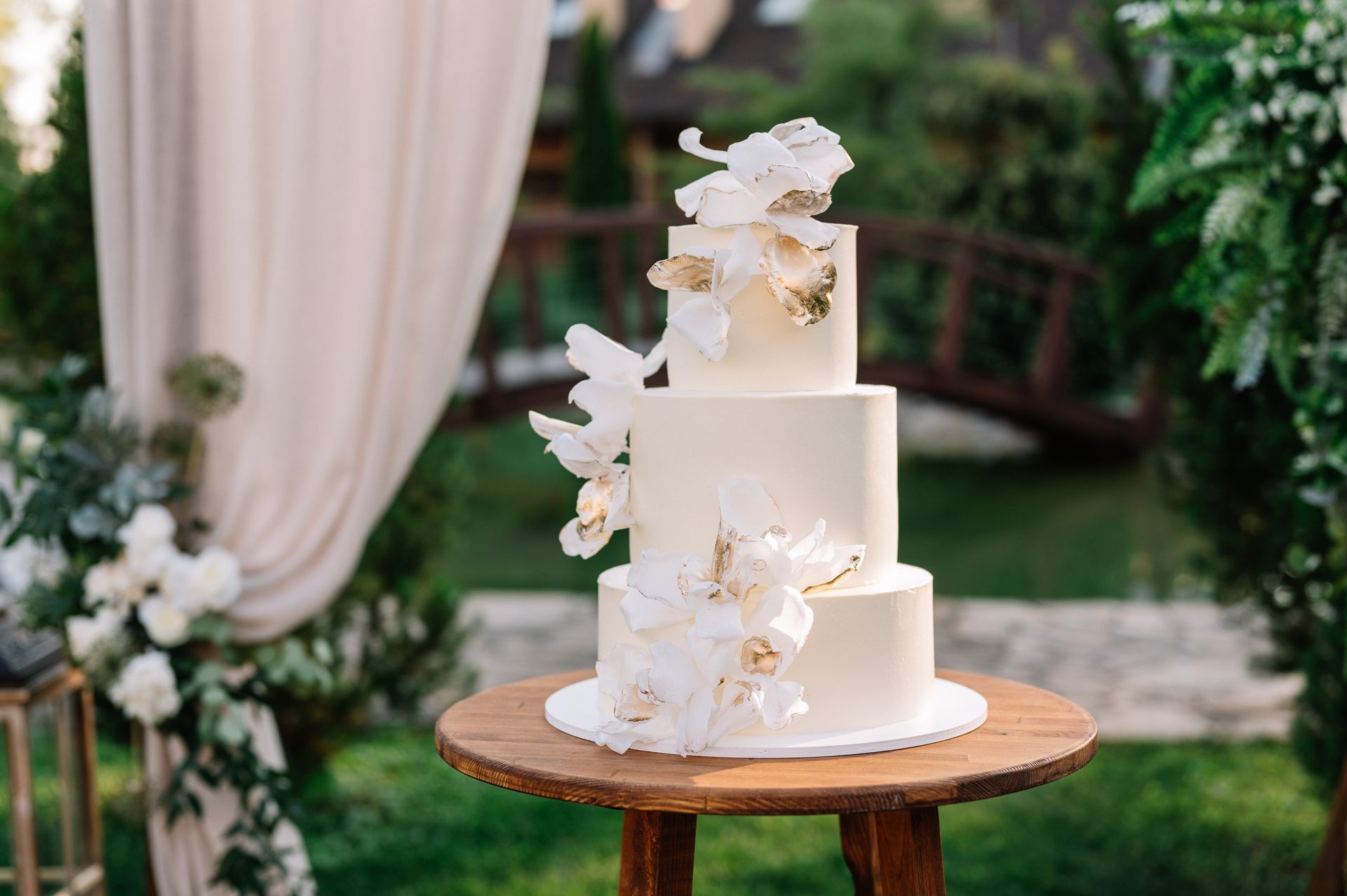 Three-tier white wedding cake with floral decorations on a wooden table in a garden setting