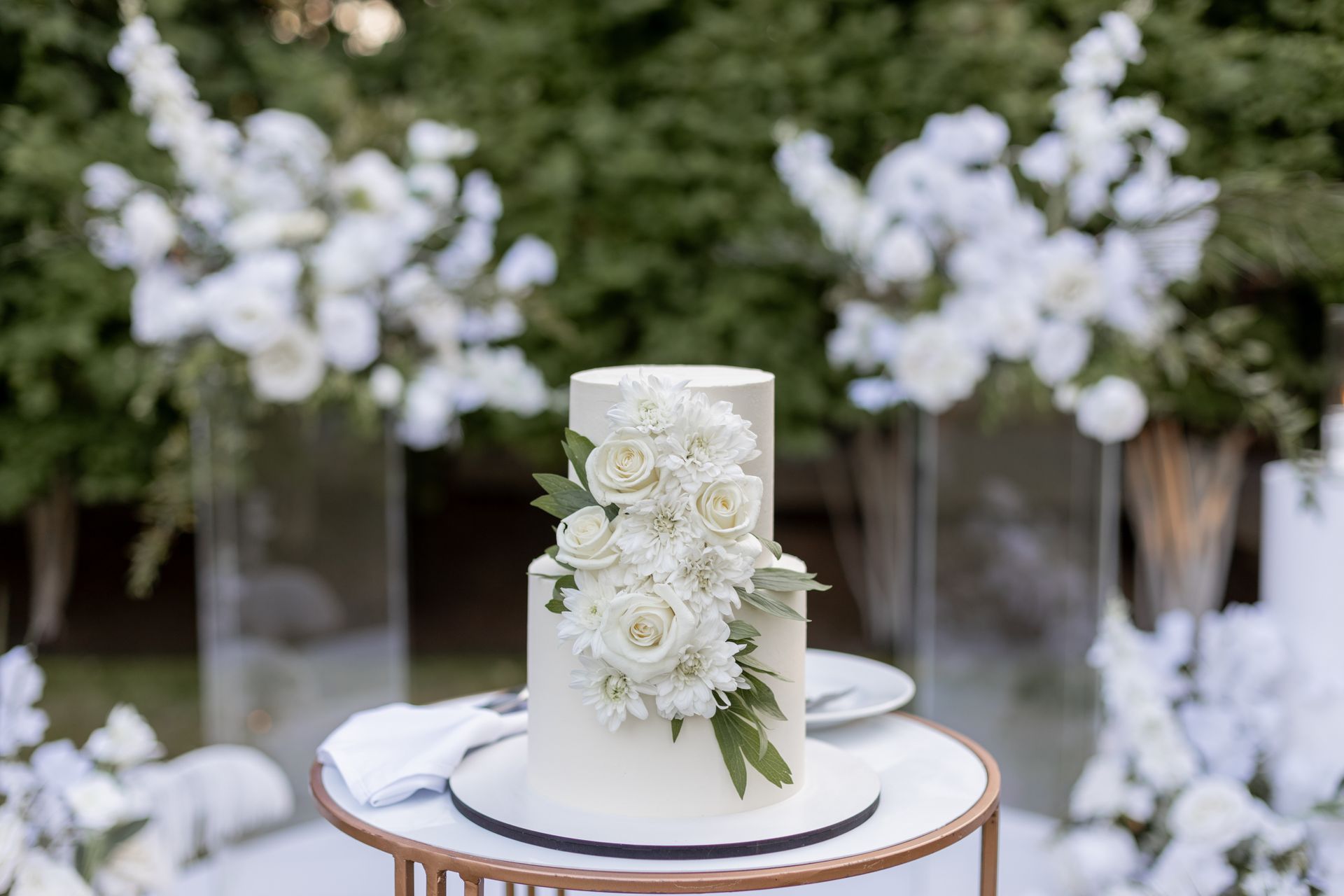 White floral wedding cake on a round table with blurred white flower arrangements in the background
