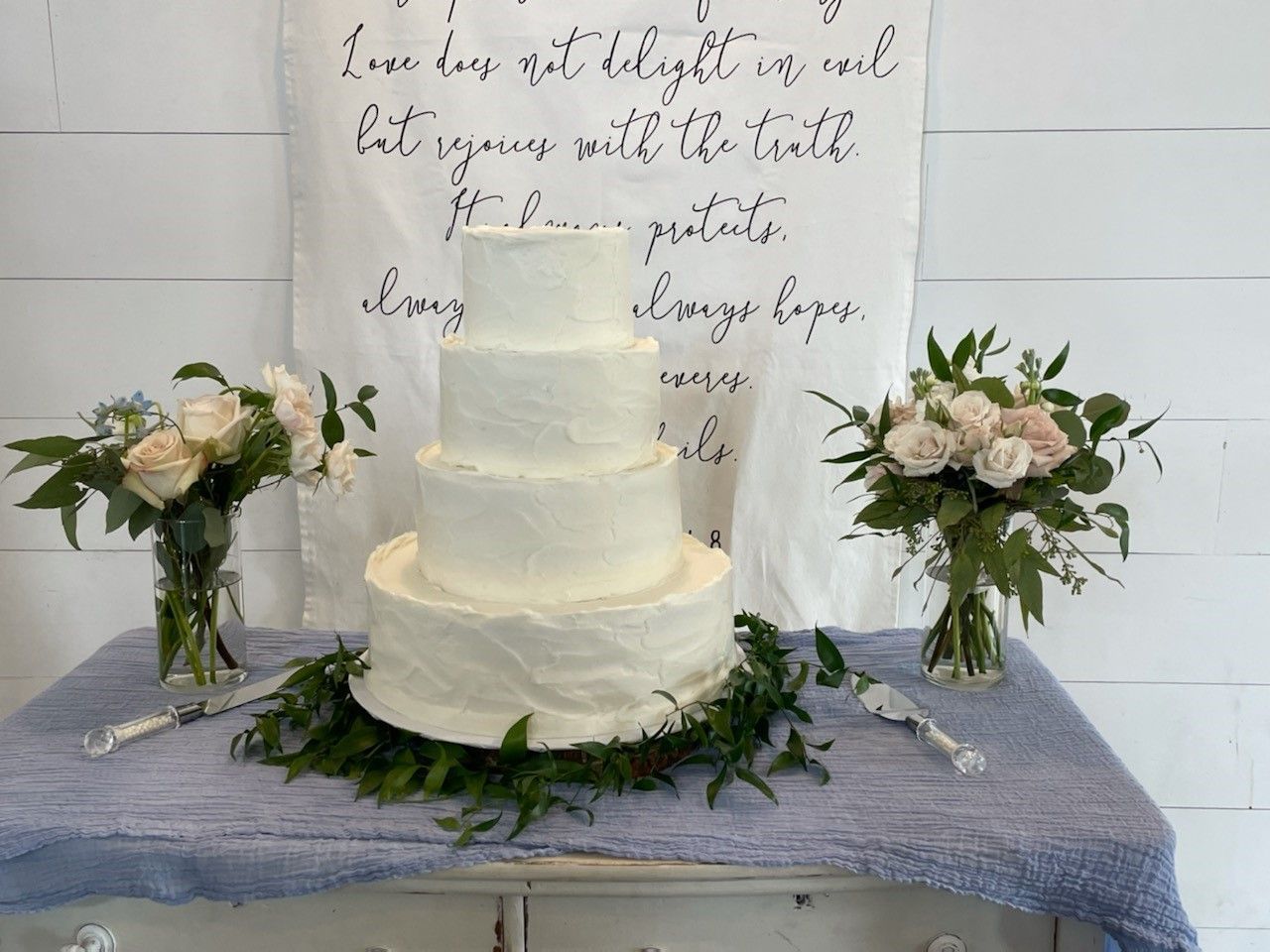 White tiered cake on a lavender tablecloth, flanked by floral arrangements in front of a handwritten sign.