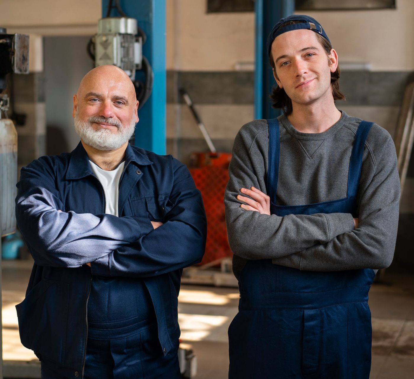Two men are standing next to each other in a garage with their arms crossed.