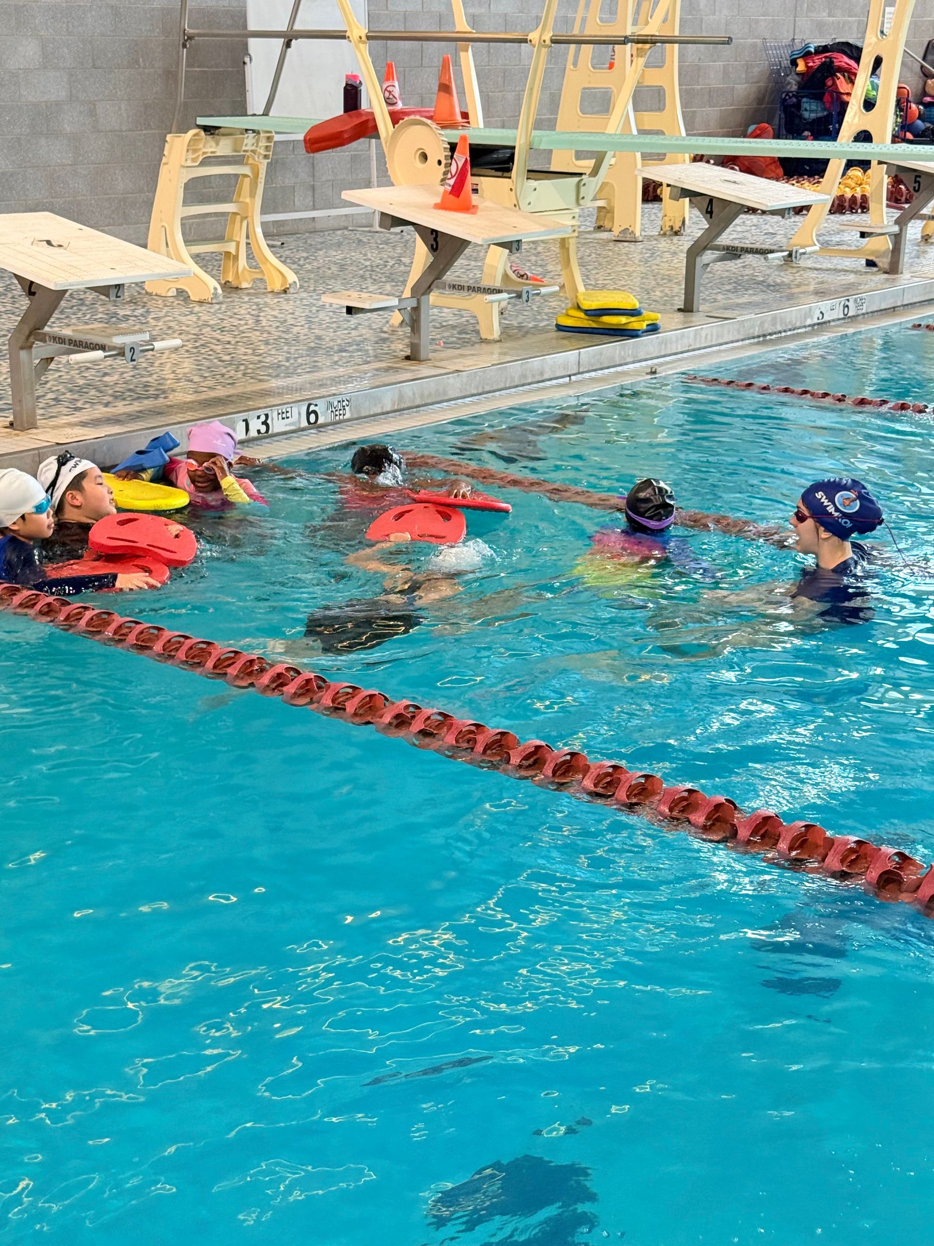 Child floating in pool, swimming instructor nearby. Blue water, orange cap.