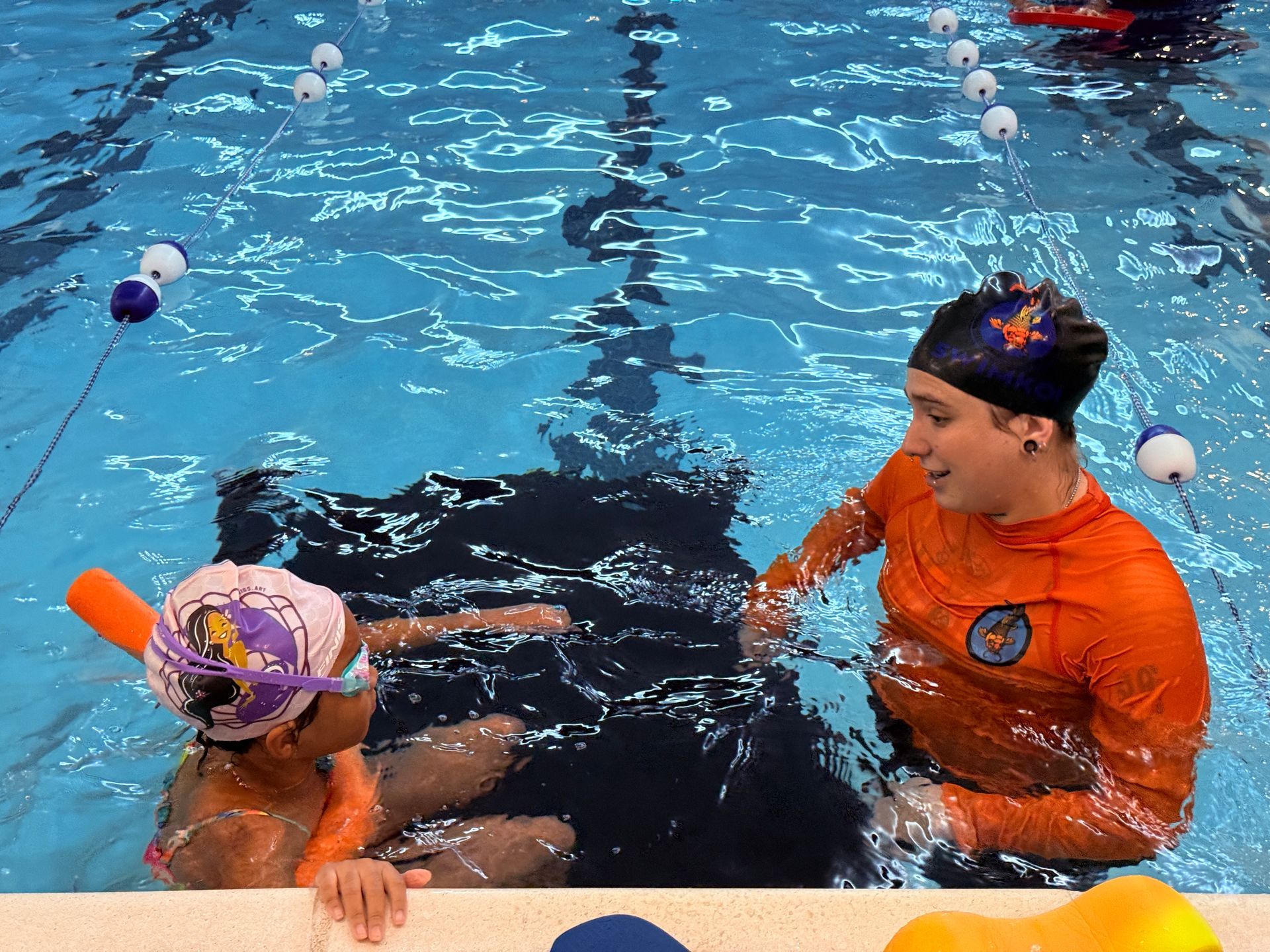 A person guides a child in a pool, teaching them to swim with a kickboard.