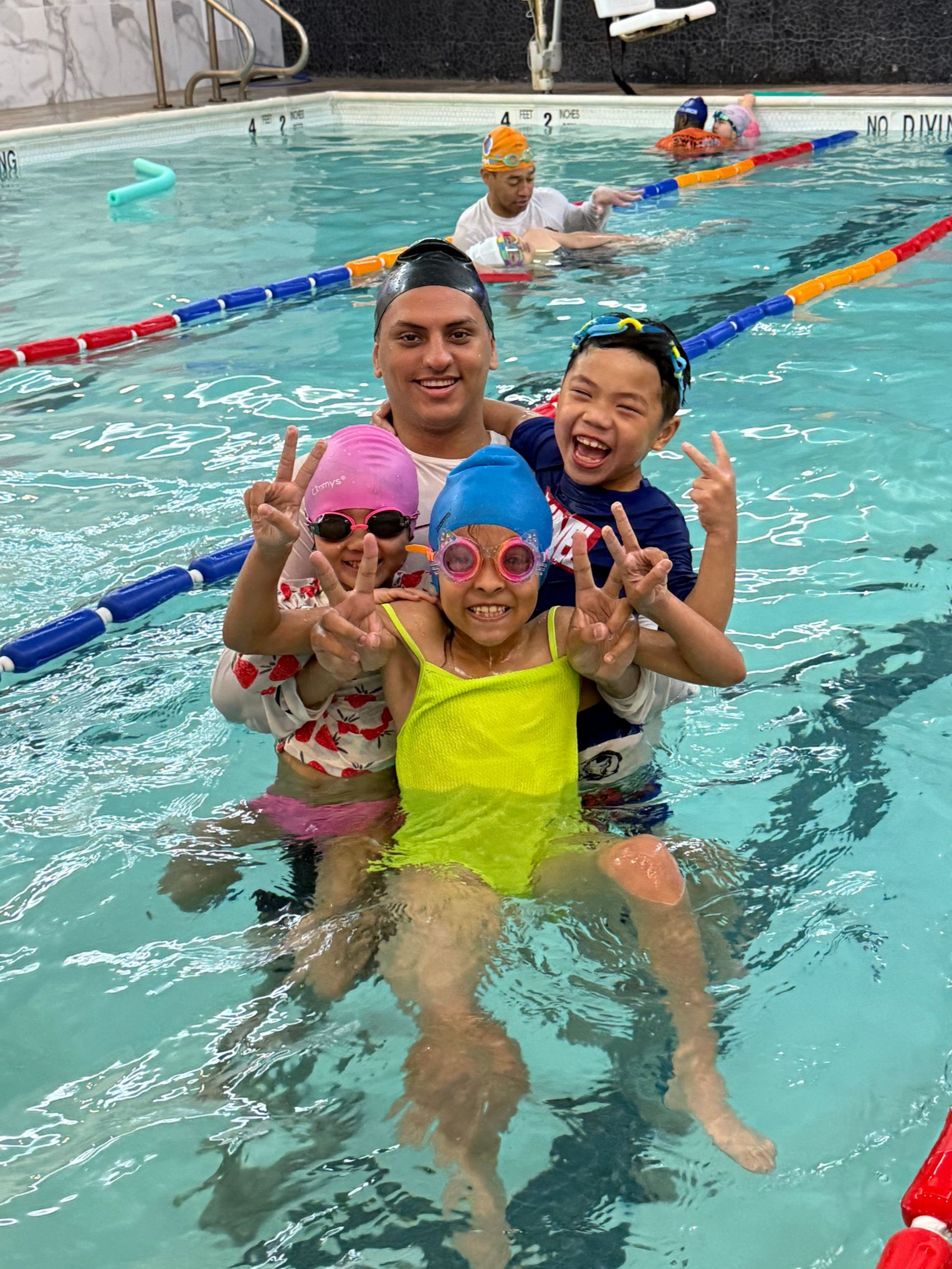 Children in swim caps and goggles in a pool, smiling and holding onto the edge.
