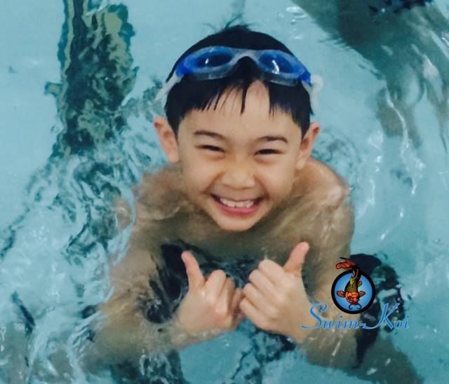 Boy with swim goggles in a pool, smiling and giving thumbs up.