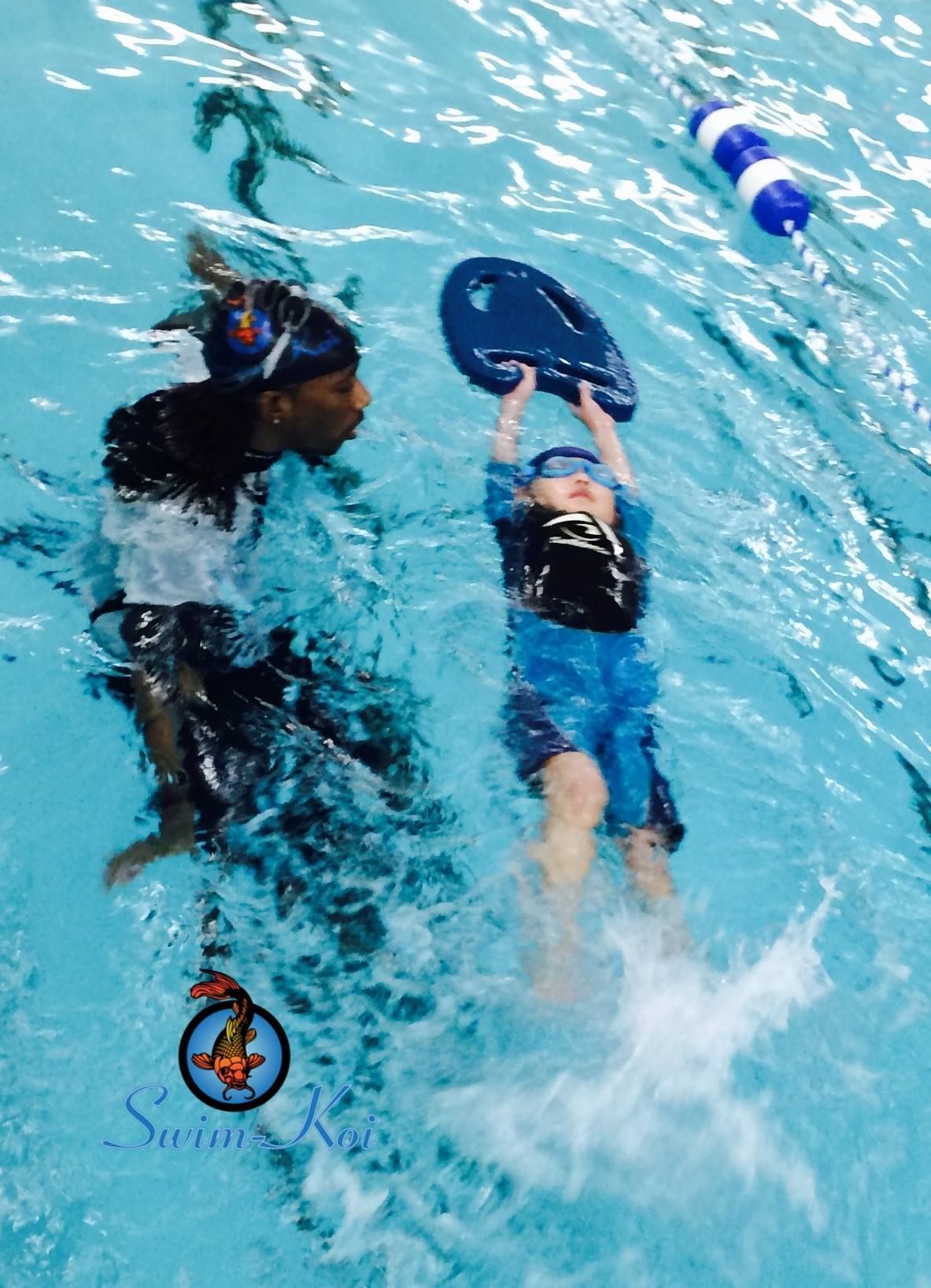A person guides a child in a pool, teaching them to swim with a kickboard.