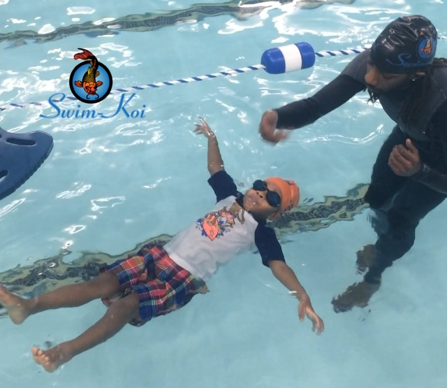 Child floating in pool, swimming instructor nearby. Blue water, orange cap.