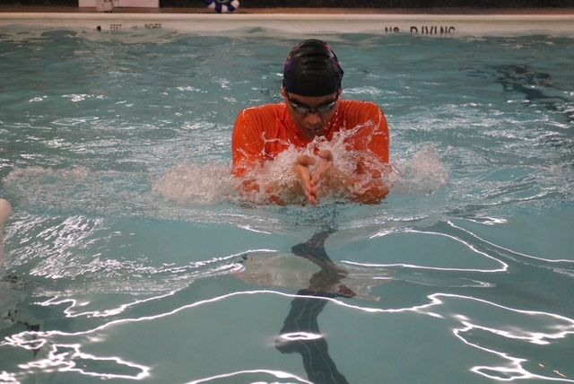 Person in orange shirt swimming breaststroke in a pool, hands creating splashing water.