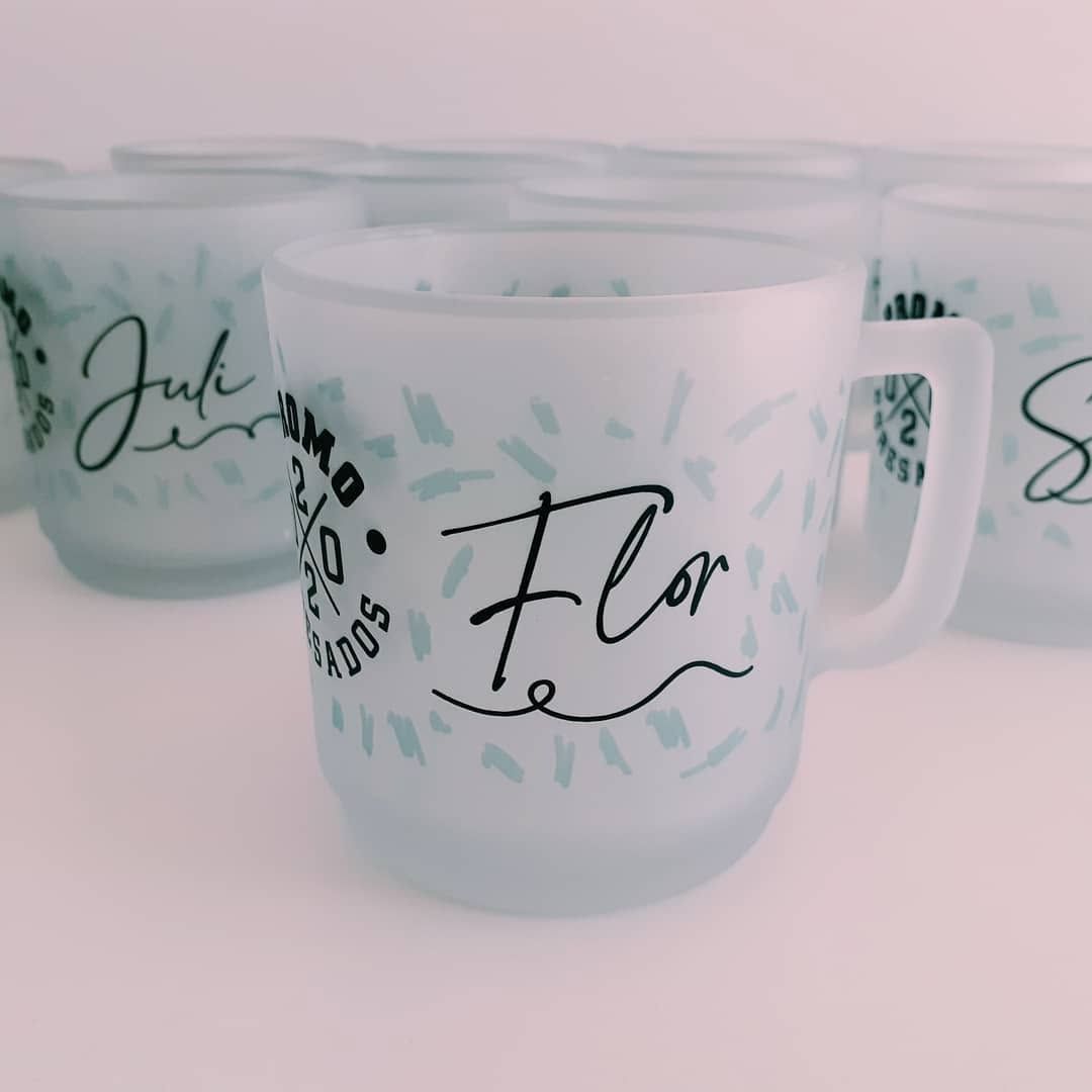 Tres tazas de vidrio esmerilado con nombres escritos en letra negra sobre un fondo rosa pálido.