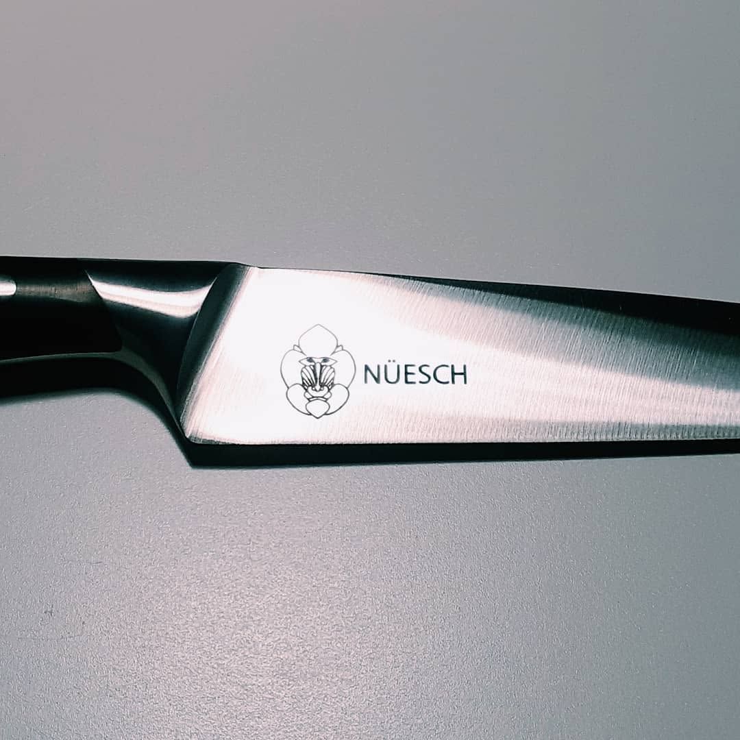 Primer plano de la hoja plateada de un cuchillo Nüesch sobre un fondo gris.