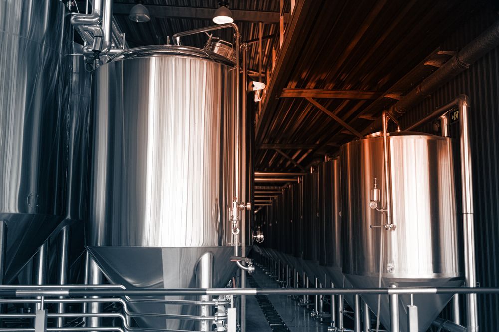 Stainless steel brewery tanks in an industrial production room with pipes and warm lighting