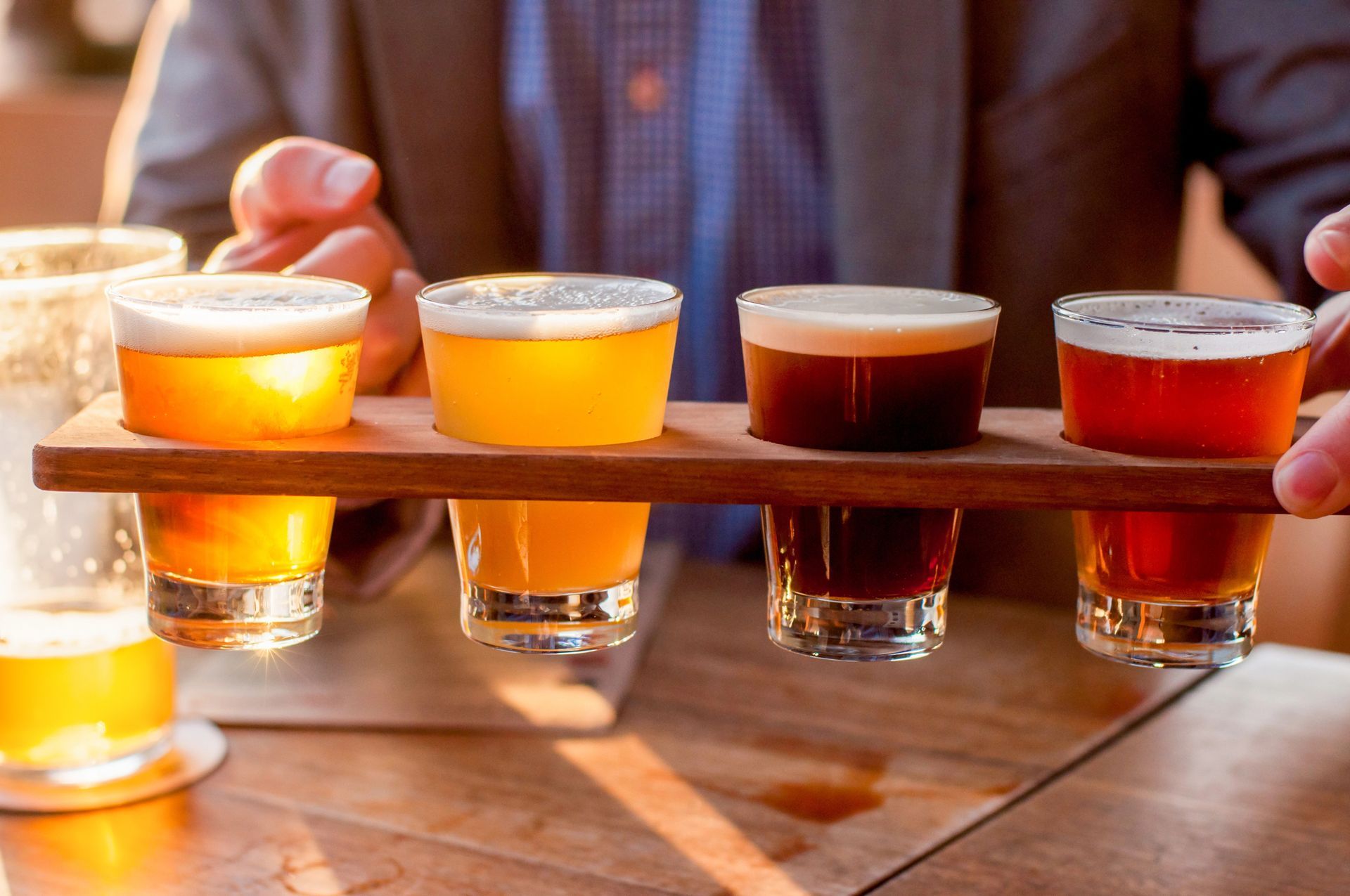 Four small beer samples on a wooden tray, with one person holding the tray outdoors in warm sunlight