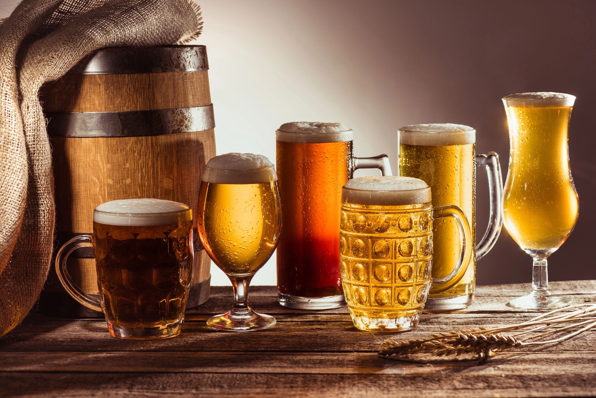 Assorted beers in glasses and mugs on a rustic table beside a wooden barrel
