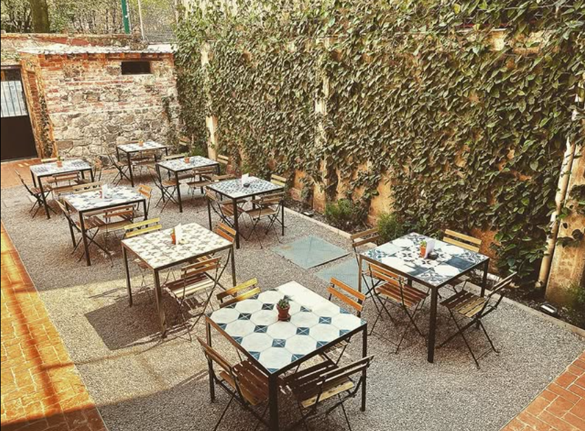 Cafetería al aire libre con mesas y sillas cuadradas en un patio de grava, con un muro cubierto de hiedra como telón de fondo.