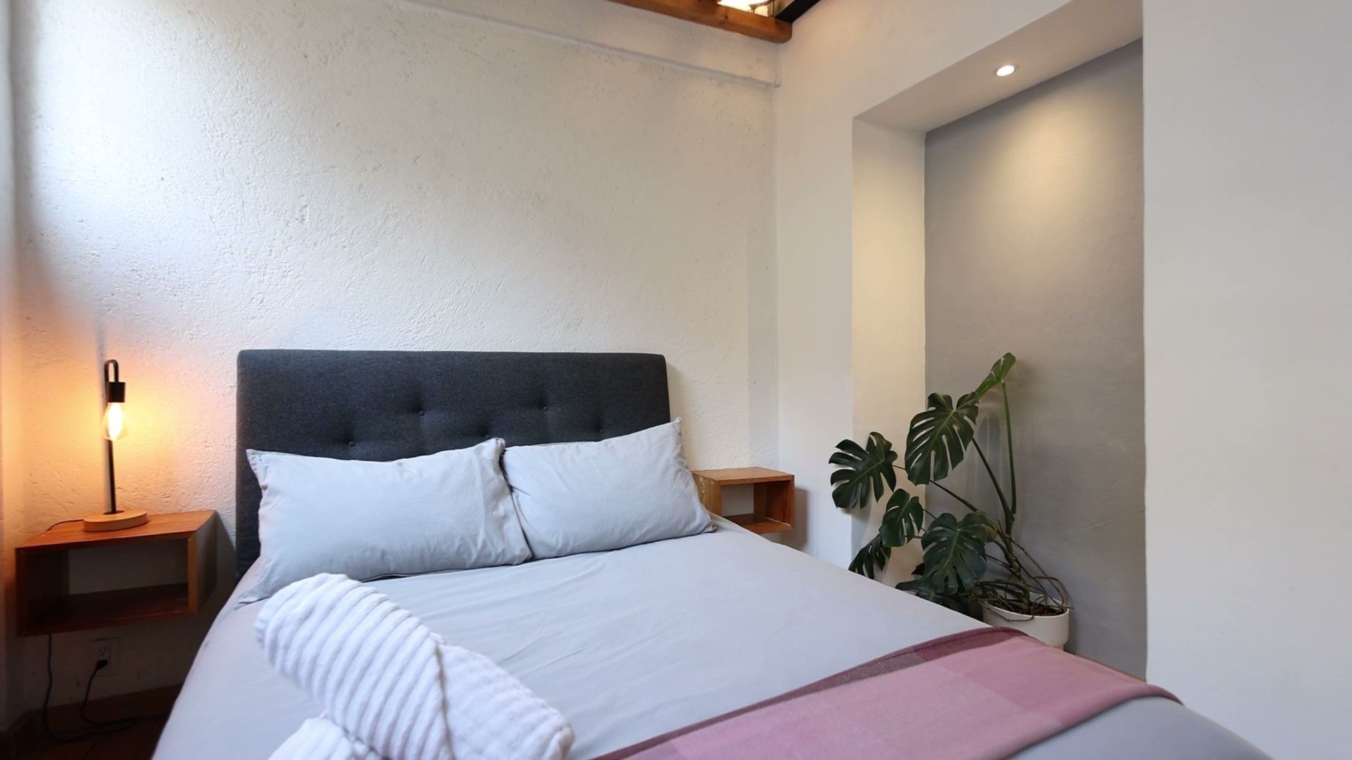 Un dormitorio con cama, mesita de noche, lámpara y planta.