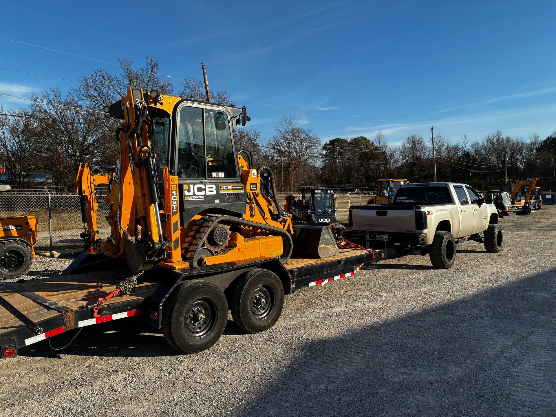 White pickup towing a trailer with a yellow backhoe on a gravel lot under a blue sky