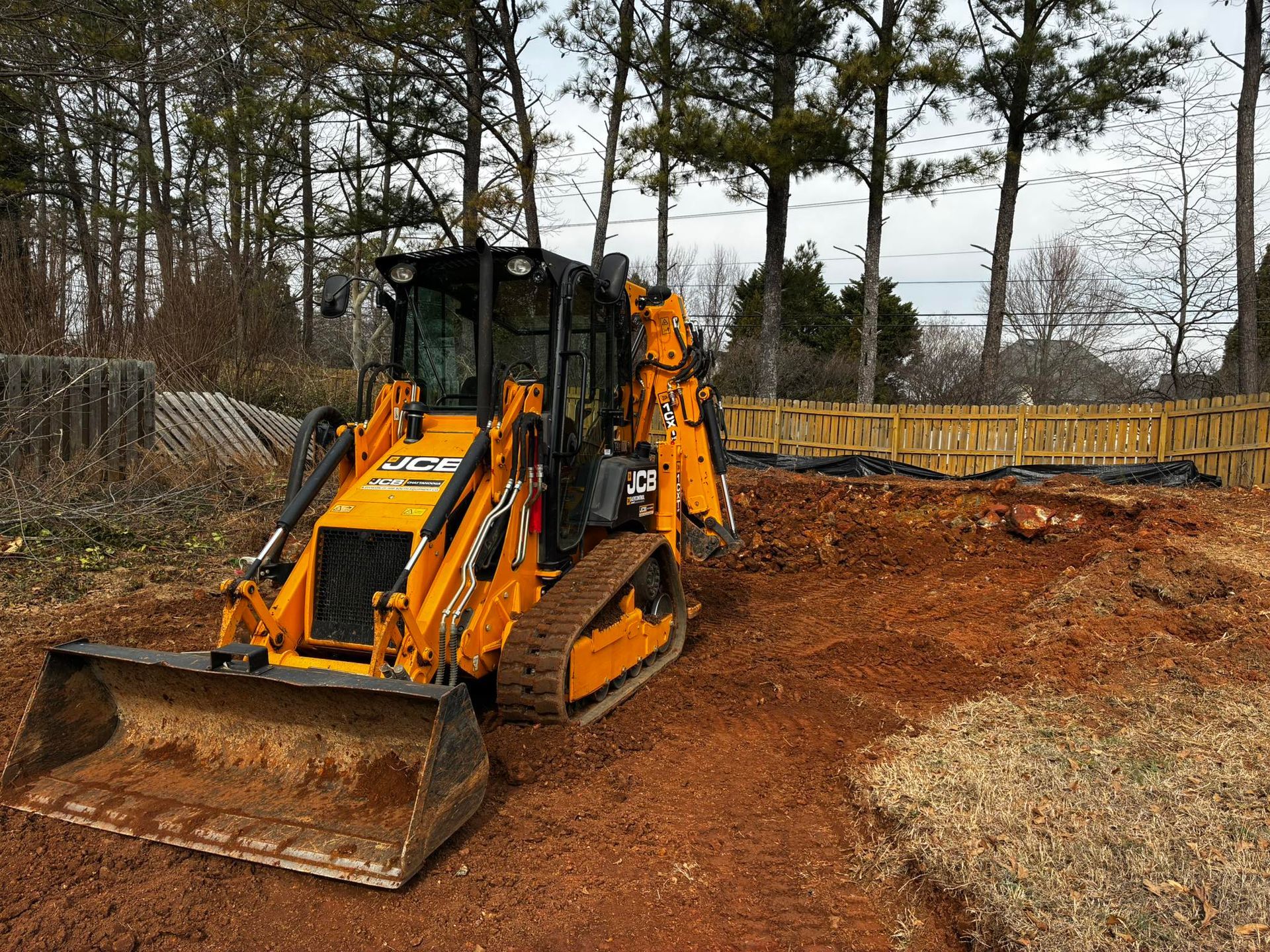 Orange backhoe loader with front bucket parked on a dirt site near trees and a wooden fence.