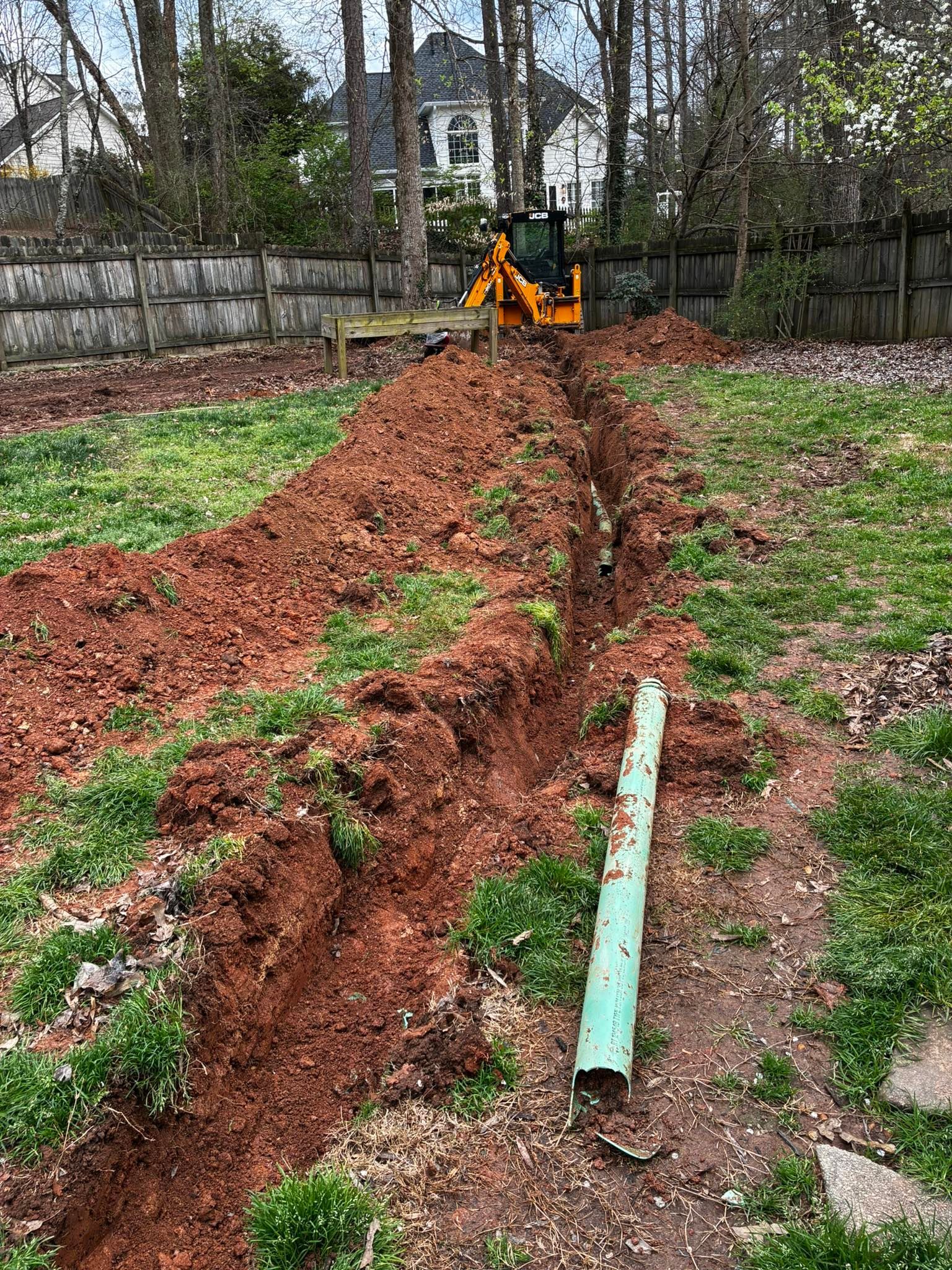 A muddy trench runs through a yard toward an orange excavator near a fence and trees.