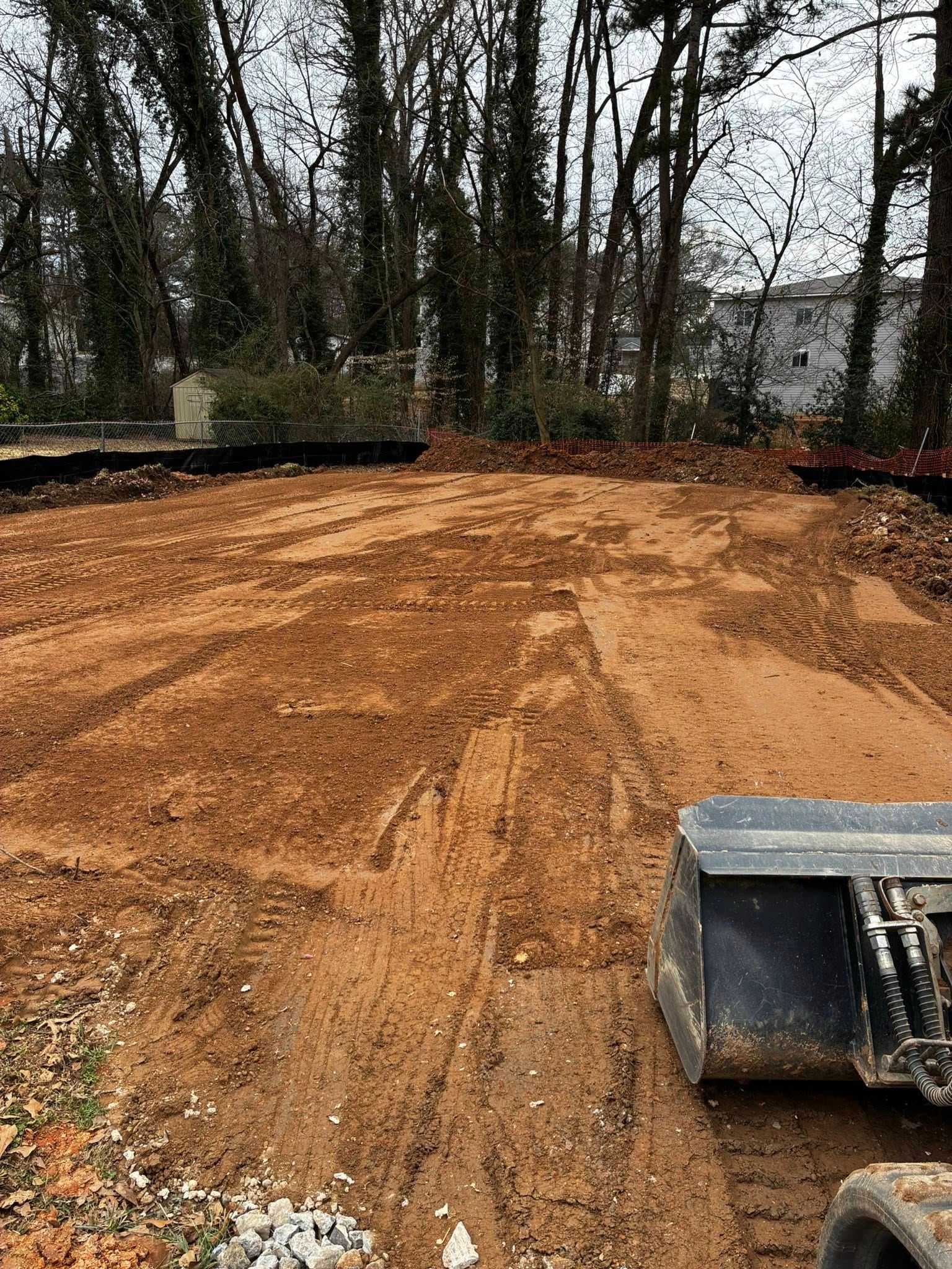 Cleared dirt lot with trees in the background and a small excavator bucket at the lower right