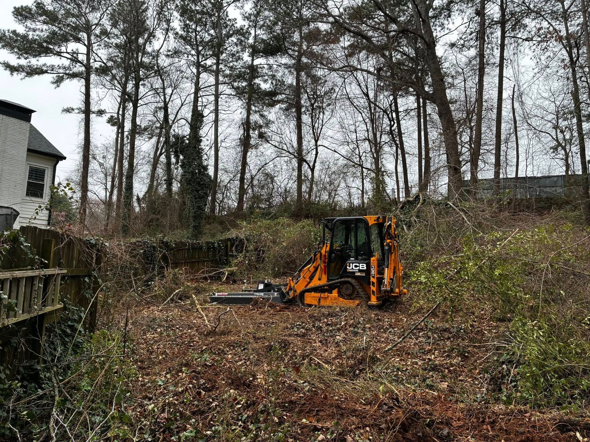 Orange excavator clearing brush on a wooded hillside near a house