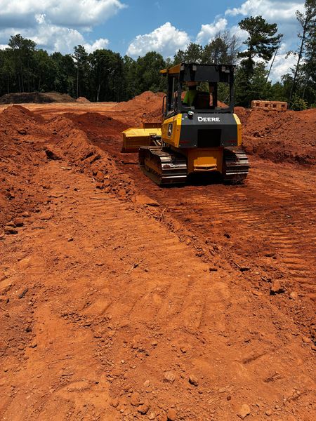 Backhoe loader digging red dirt, with operator in a yellow vest at a wooded work site