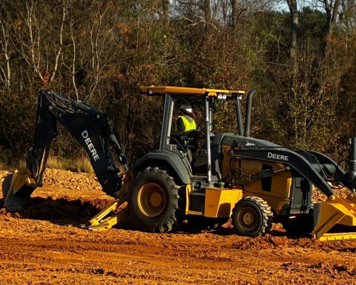 Backhoe loader digging orange soil at a construction site, with a worker in the cab